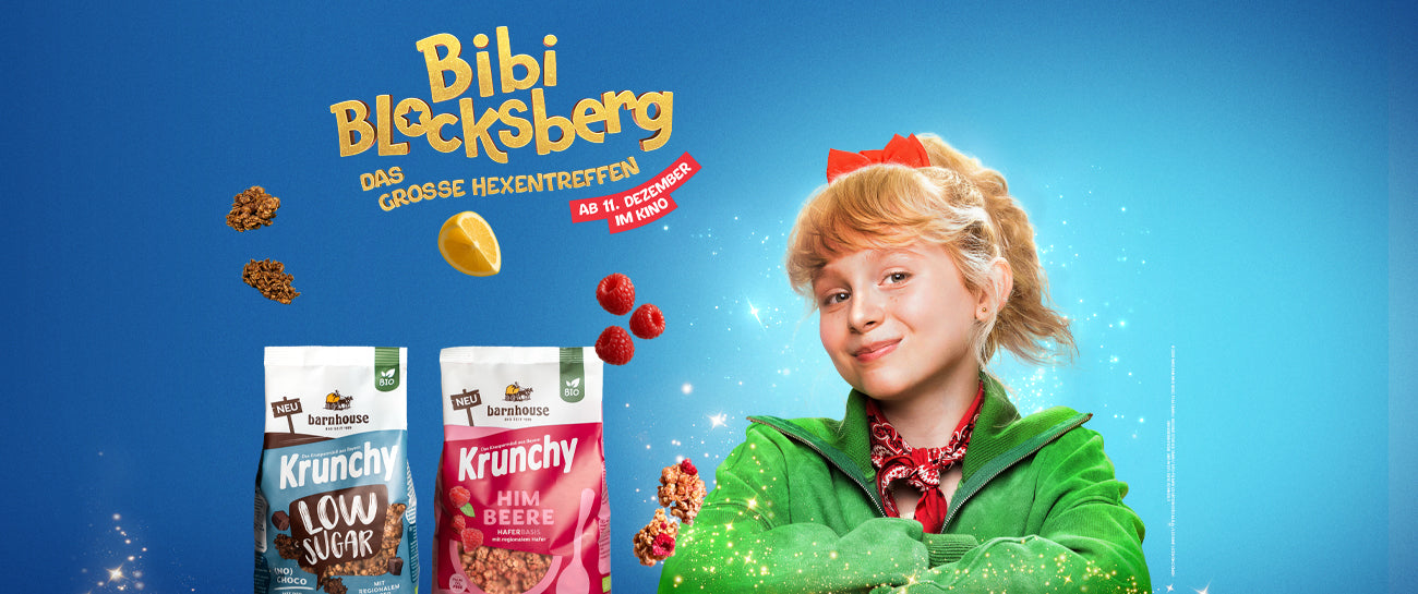 Blondes, junges Mädchen mit grüner Jacke und roter Schleife. Außenrum Glitzer. Daneben 2 Krunchy-Beutel, Cluster, Himbeeren & 1 Zitrone. Links oben steht "Bibi Blocksberg - DAS GROSSE HEXENTREFFEN ab 11. Dezember im Kino".