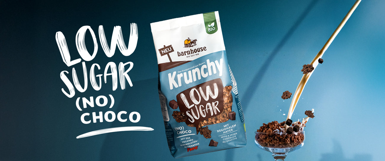 Beutel Krunchy Low Sugar (No) Choco neben Glas mit Knuspermüsli. Milchstrahl fließt ins Glas, Schoko-Cluster fliegen, links der Schriftzug Low Sugar (No) Choco.