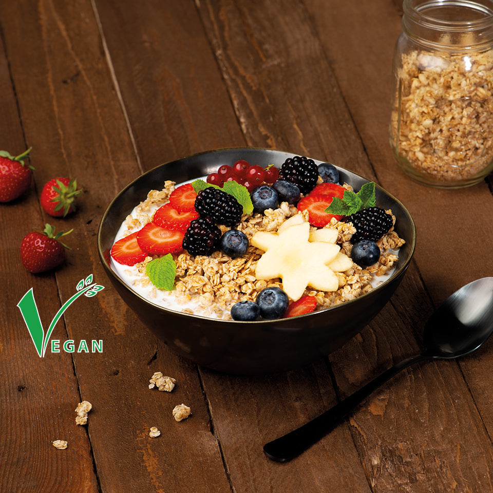 Sehr schön angerichtete Bowl mit Krunchy, frischen Brombeeren, Erdbeerscheiben, Johannisbeeren, Heidelbeeren, Sternenfrucht und etwas Minze. Auf dem Holztisch liegen auch frische Erdbeeren, ein paar Müslicluster und ein Löffel.