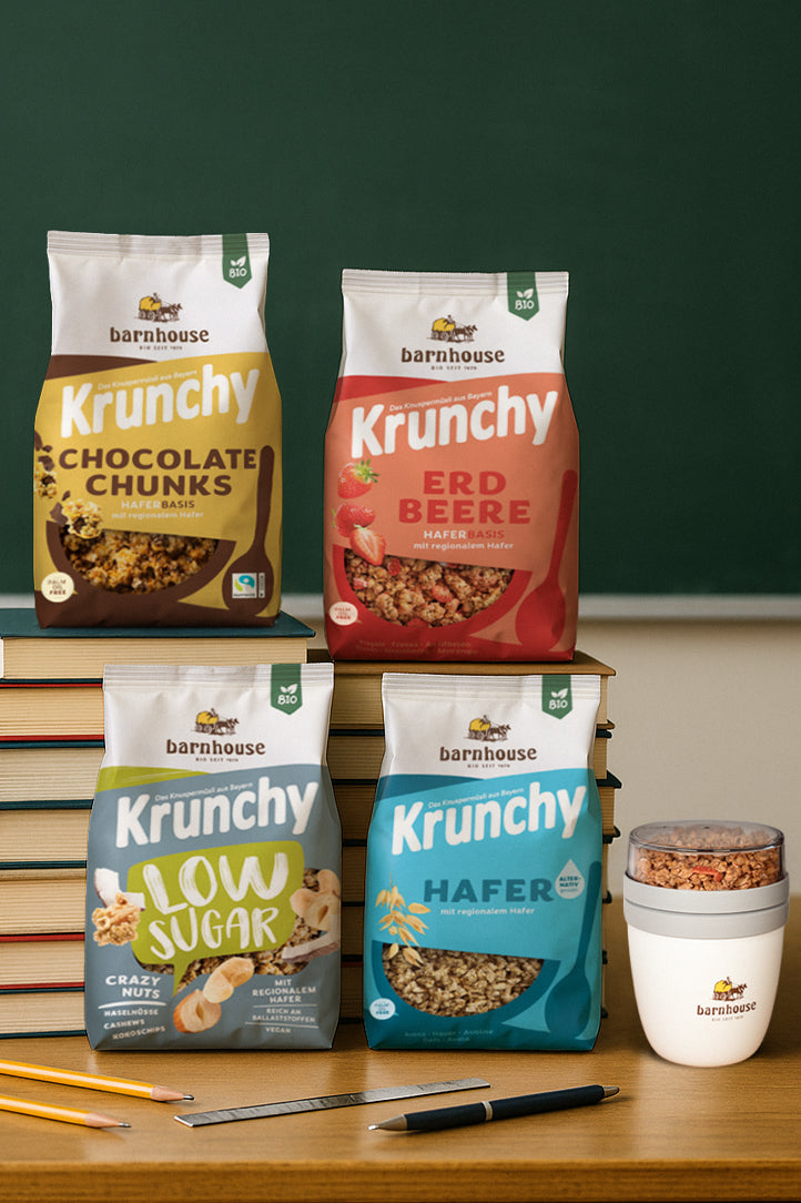 Holztisch, der vor einer grünen Tafel im Klassenzimmer steht. Auf dem Tisch liegen 2 Bleistifte, 1 Kugelschreiber und 1 Lineal. Dahinter stehen je eine XL-Packung Krunchy Low Sugar Crazy Nuts und Hafer alternativ gesüßt. Dahinter sind 2 Türme mit gestapelten Büchern, auf denen eine XL-Packung Krunchy Chocolate Chunks und Erdbeere stehen. Auf der rechten Seite steht ein befüllter Krunchy2go-Becher.