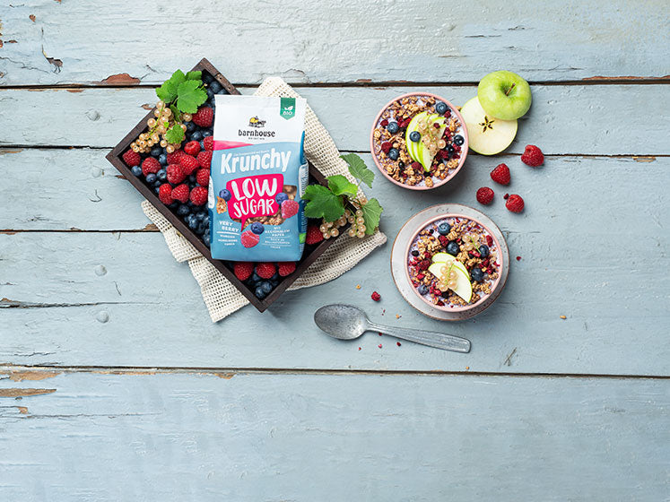 Blick von oben auf einen rosa-weiß-blauen Beutel Krunchy Low Sugar Very Berry von Bio-Knuspermüsli-Hersteller Barnhouse, der in einer Kiste mit Himbeeren, Heidelbeeren und weißen Johannisbeeren auf einer Decke liegt. Daneben stehen 2 rosa Schüsseln (eine mit Unterteller) mit Milch & diesem zuckerarmen Müsli - jeweils mit Himbeeren, Heidelbeeren, weißen Johannisbeeren und grünen Apfelscheiben dekoriert. Auf dem bläulich gestrichenen Holztisch liegen ein silberner Löffel, ein geschnittener Apfel & Himbeeren.