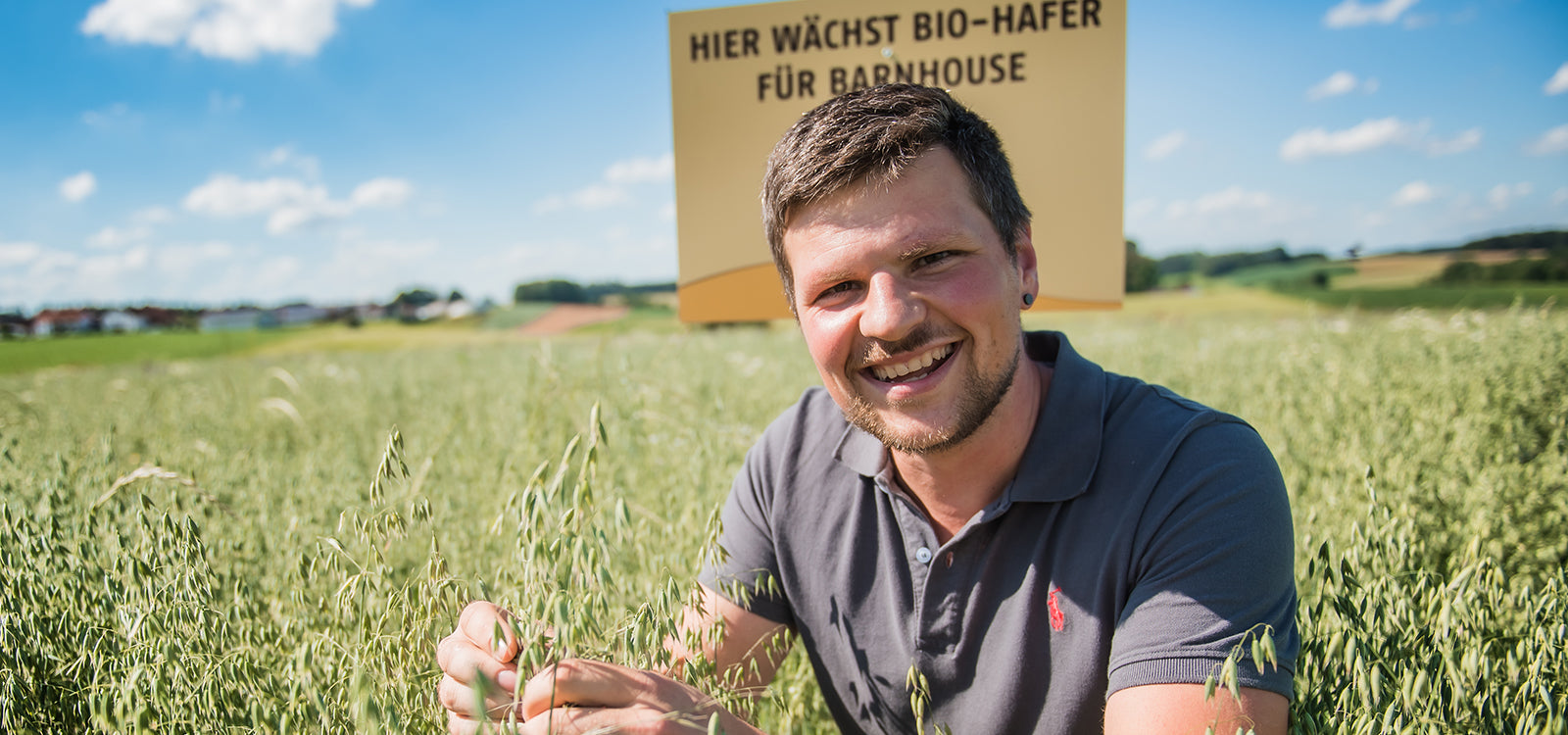 Barnhouse Bauer Johannes Hobmaier, der mit einem breiten Grinsen in seinem Bio-Haferfeld sitzt. Hinter ihm steht ein Schild mit der Aufschrift "HIER WÄCHST BIO-HAFER FÜR BARNHOSUE" und im Hintergrund ist eine tolle Landschaft zu sehen.