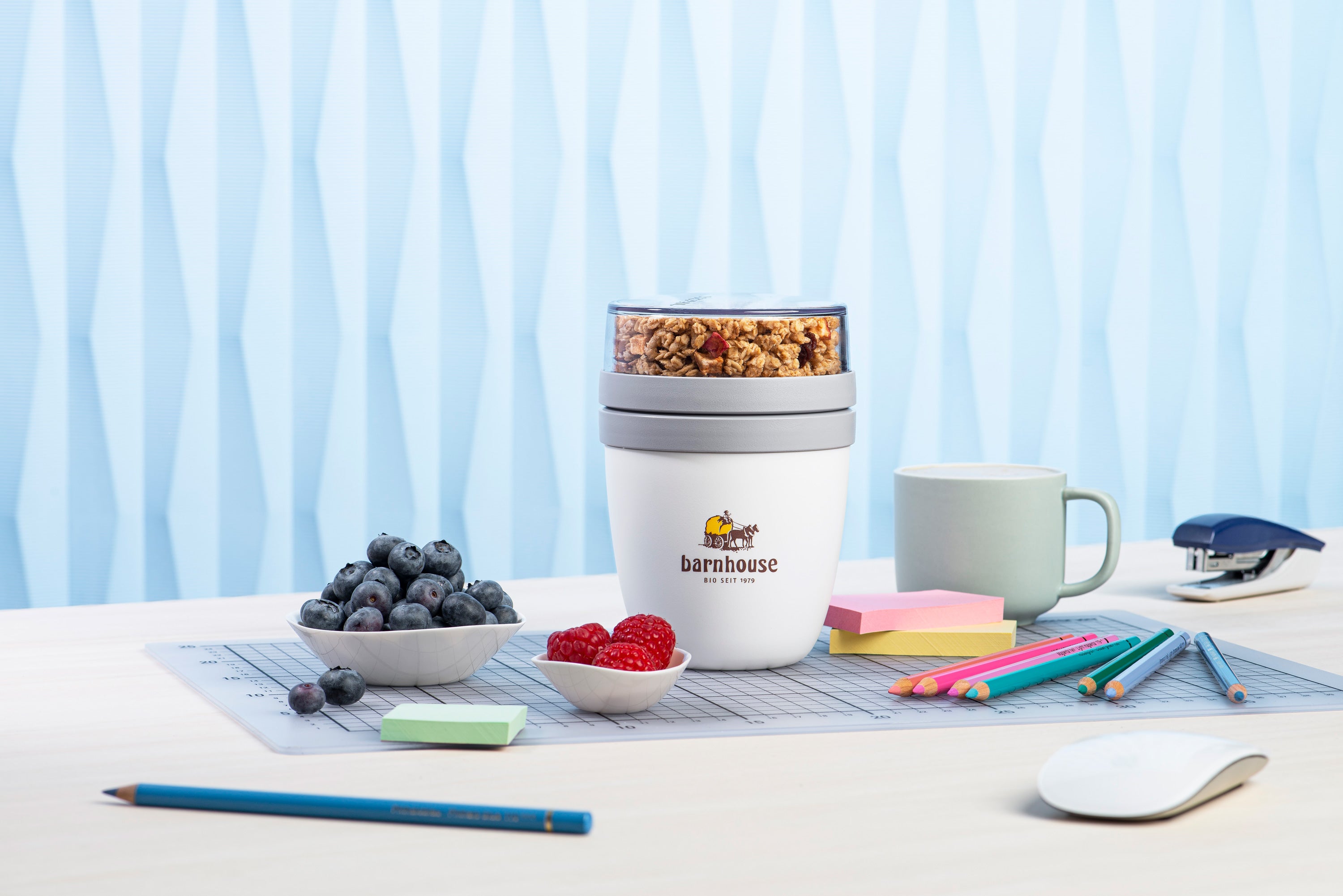 Blick von vorne auf einen Bürotisch. Zwischen den Büromaterialien Bleistift, Computermaus, Post-its, Farbstiften und Tacker steht ein Krunchy2go-Becher von Bio-Knuspermüsli-Hersteller Barnhouse auf der Schreibtisch-Unterlage. Daneben stehen noch eine Tasse sowie je eine weiße Schale mit Heidelbeeren und Himbeeren.