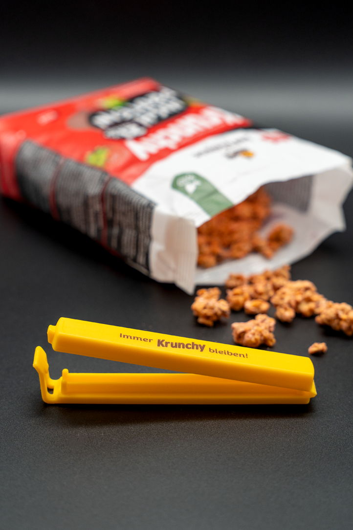 Der Fokus liegt auf dem gelben Beutelclip des Bio-Knuspermüsli-Herstellers Barnhouse mit der braunen Aufschrift "Immer Krunchy bleiben". Im Hintergrund liegt eine offene Packung Krunchy High Protein, aus der ein paar Cluster in Richtung Clip rutschen.