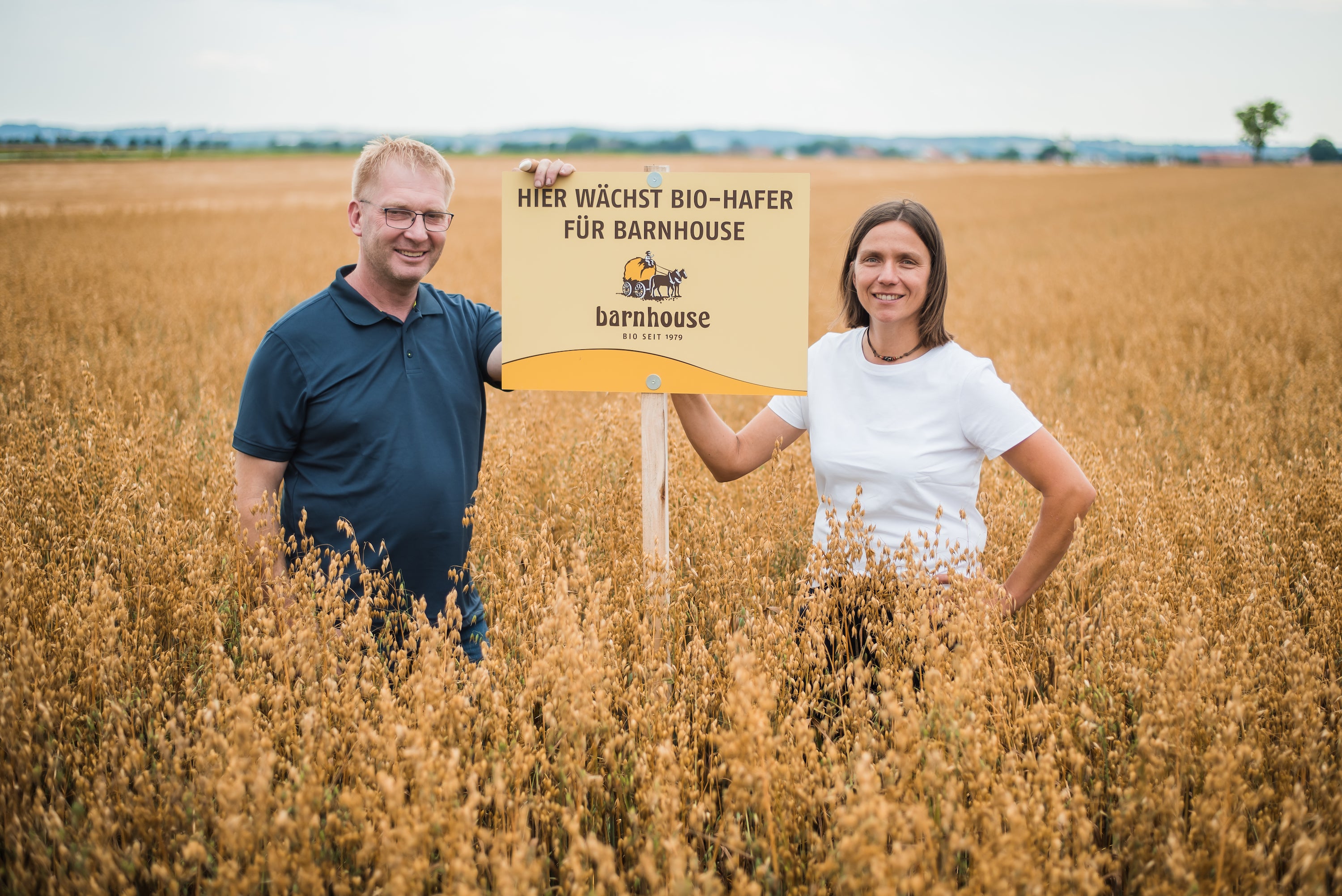 Susanne Fischer und ihr Mann in ihrem Bio-Haferfeld. Beide halten das Schild mit der Aufschrift "HIER WÄCHST BIO-HAFER FÜR BARNHOUSE" fest.