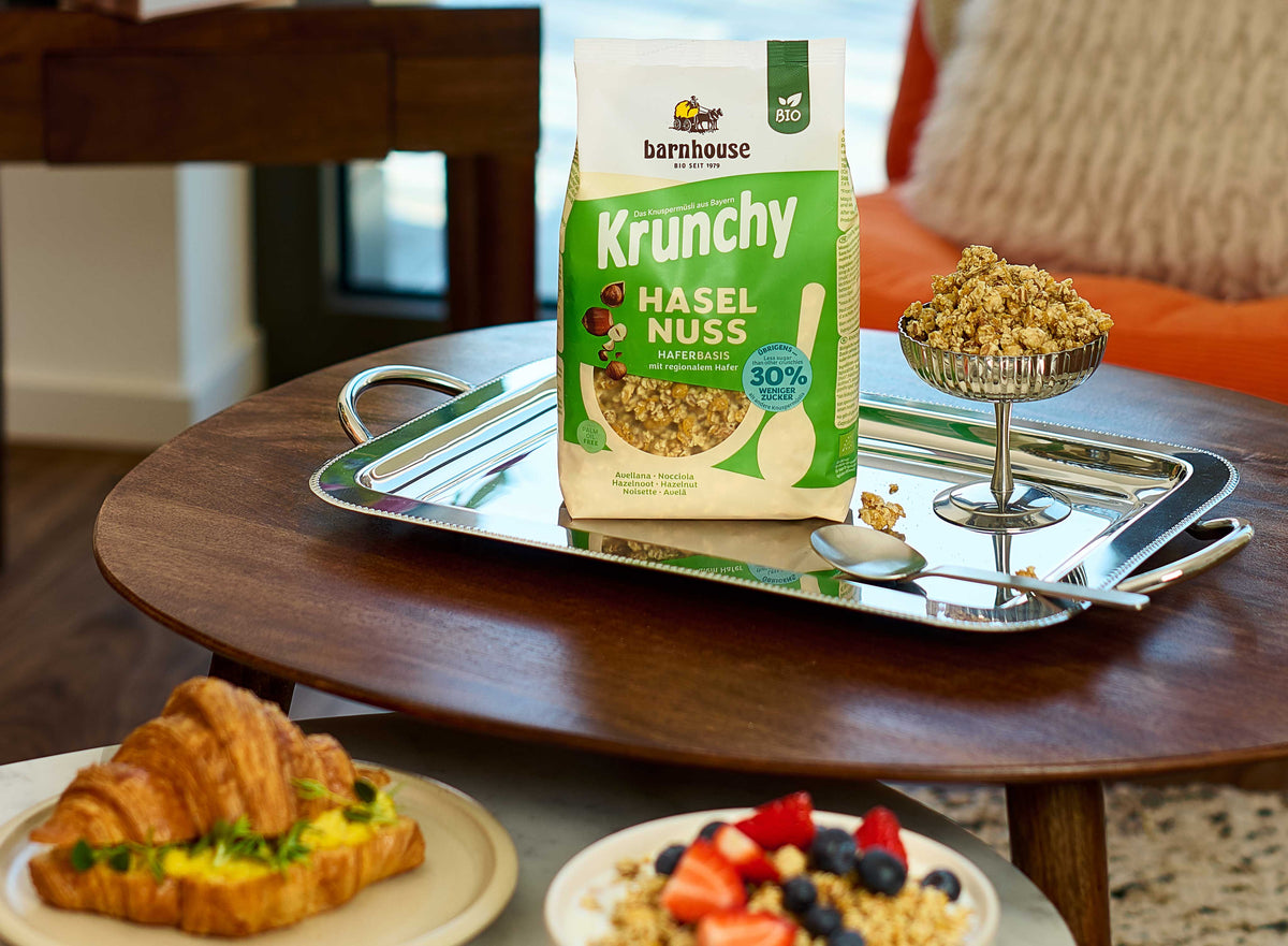 Blick von vorne auf einen grün-beigen Beutel Krunchy Haselnuss weniger Zucker von Barnhouse, der auf einem silbernen Tablett steht. Dort liegen außerdem ein Müsli-Cluster, ein silberner Löffel sowie ein mit Krunchy Haselnuss befülltes Martini-Glas. Die Sachen stehen auf einem Holztisch. Daneben ein weiterer Tisch mit einem Croissant auf einem Teller sowie eine Müslischüssel mit Krunchy Haselnuss und verschiedenen Beeren. Im Hintergrund sind ein Teppich sowie ein oranger Sessel mit beigem Kissen zu sehen.