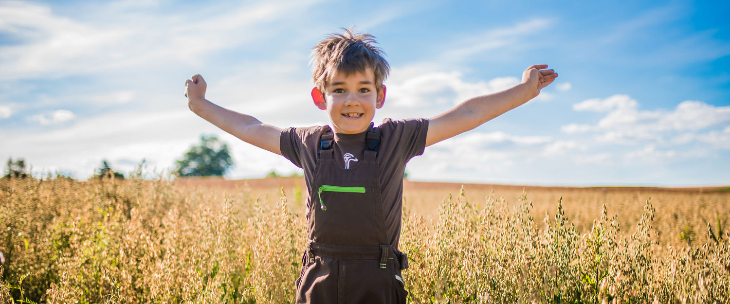 Kleiner, braunhaariger Junge, der mit braunem Engelbert Strauss T-Shirt und brauner Latzhose im Bio-Haferfeld steht und lachend seine Arme nach oben reißt. Im Hintergrund sieht man den strahlend blauen Himmel.
