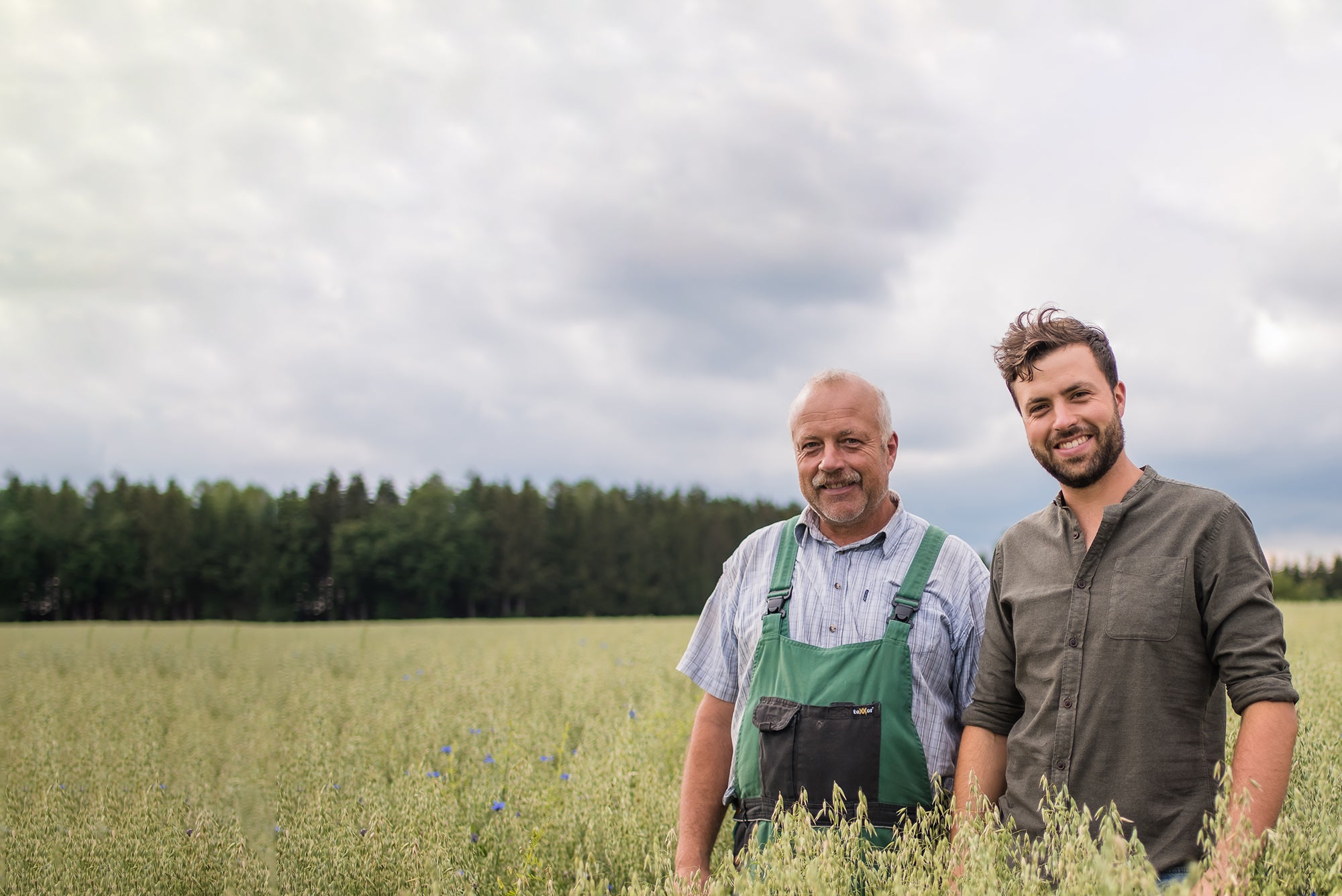 Barnhouse Bauer Alois Meier in kurzem, karierten Hemd mit grün-schwarzer Latzhose neben seinem Sohn im langen Hemd. Die beiden stehen in ihrem Bio-Haferfeld. Im Hintergrund sind ein Wald und der blaue Himmel zu sehen.