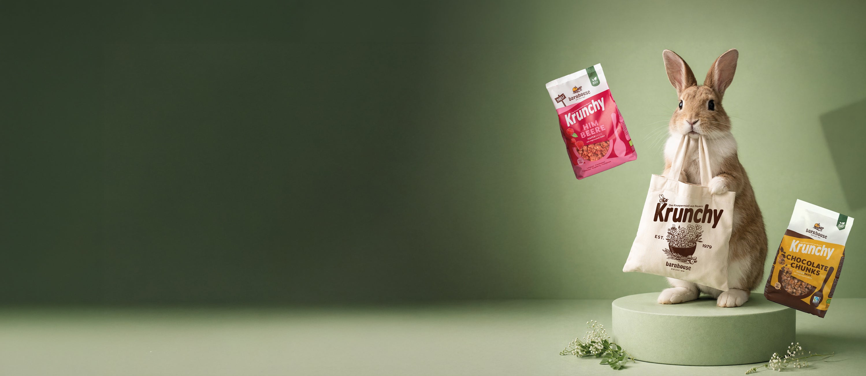 Süßer, braun-weißer Hase, der auf einem grünen Podest steht und mit seinem Mund einen Krunchy Stoffbeutel hält. Neben ihm je ein Beutel Krunchy Himbeere und Chocolate Chunks.