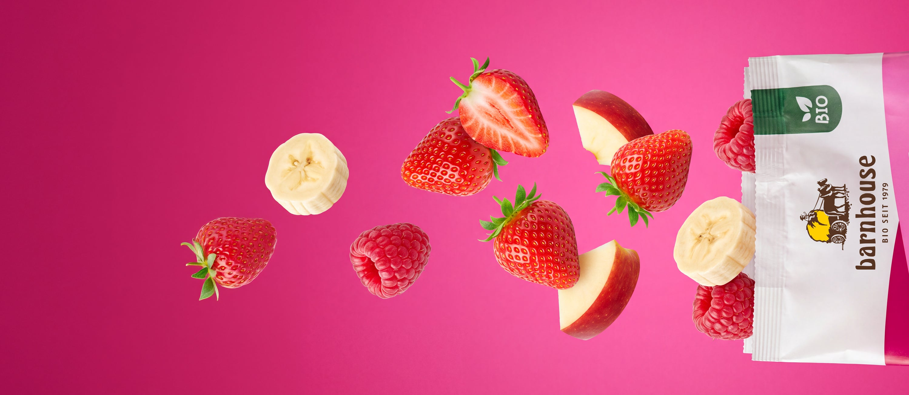 Geöffneter Beutel von Krunchy Himbeere, aus dem Himbeeren, Erdbeeren, Banananscheiben und Apfelstücke herausfliegen. Der Hintergrund hat einen rosafarbenen Verlauf.