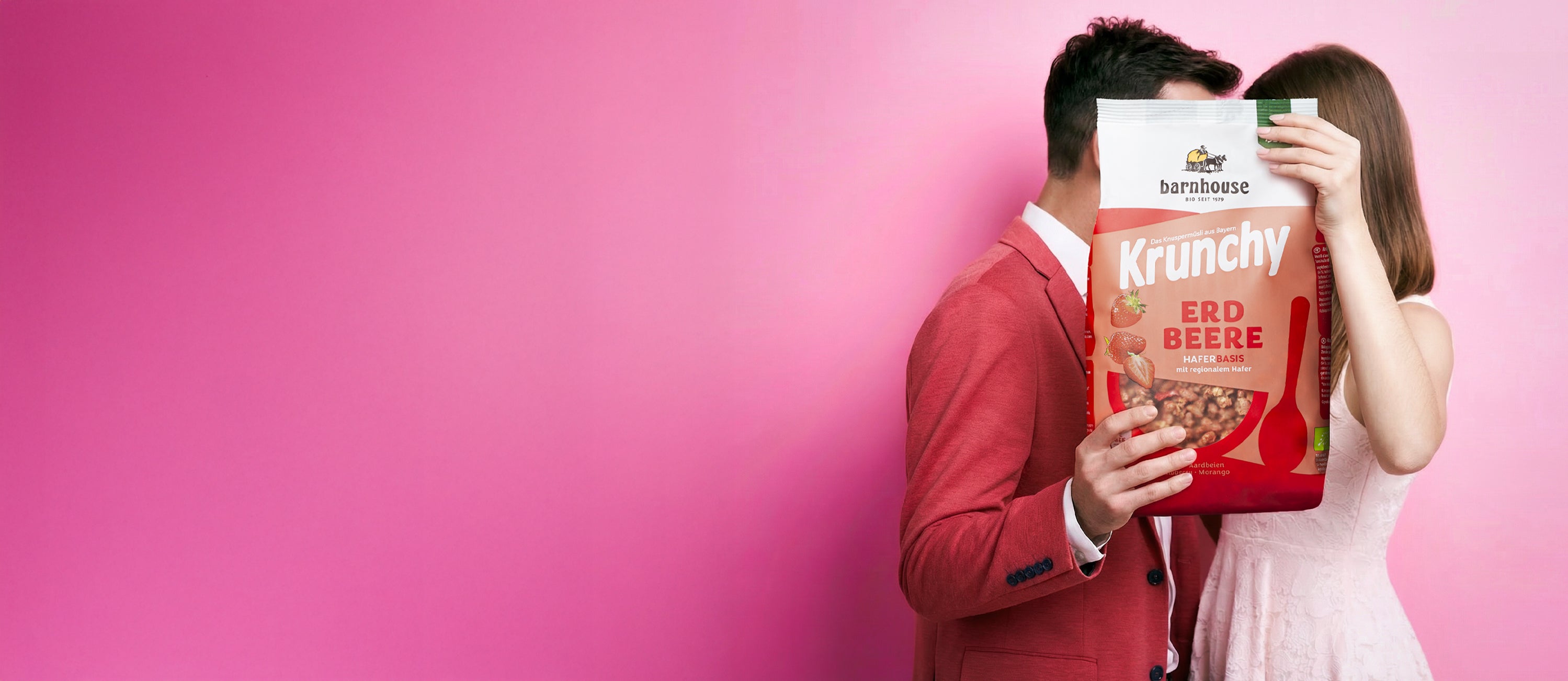 Frau im weißen Kleid & Mann im roten Anzug sind sich sehr nahe. Sieht aus, als würden sie sich küssen. Die Münder werden von XL-Packung Krunchy Erdbeere verdeckt.