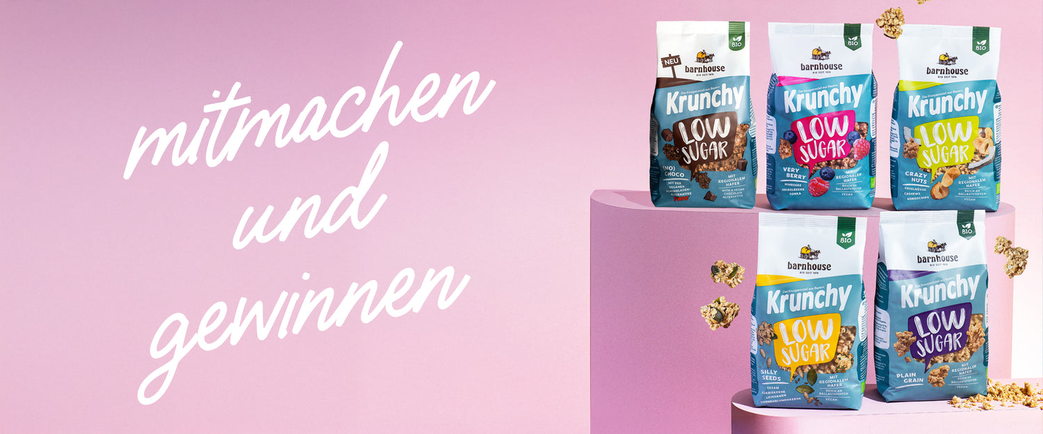 Blick von vorne auf die die fünf Low Sugar Krunchys Plain Grain, Crazy Nuts, Very Berry (No) Choco und Silly Seeds, die auf zwei verschiedenen rosa Sockeln stehen. Außenrum fliegen Cluster, links steht "mitmachen und gewinnen"