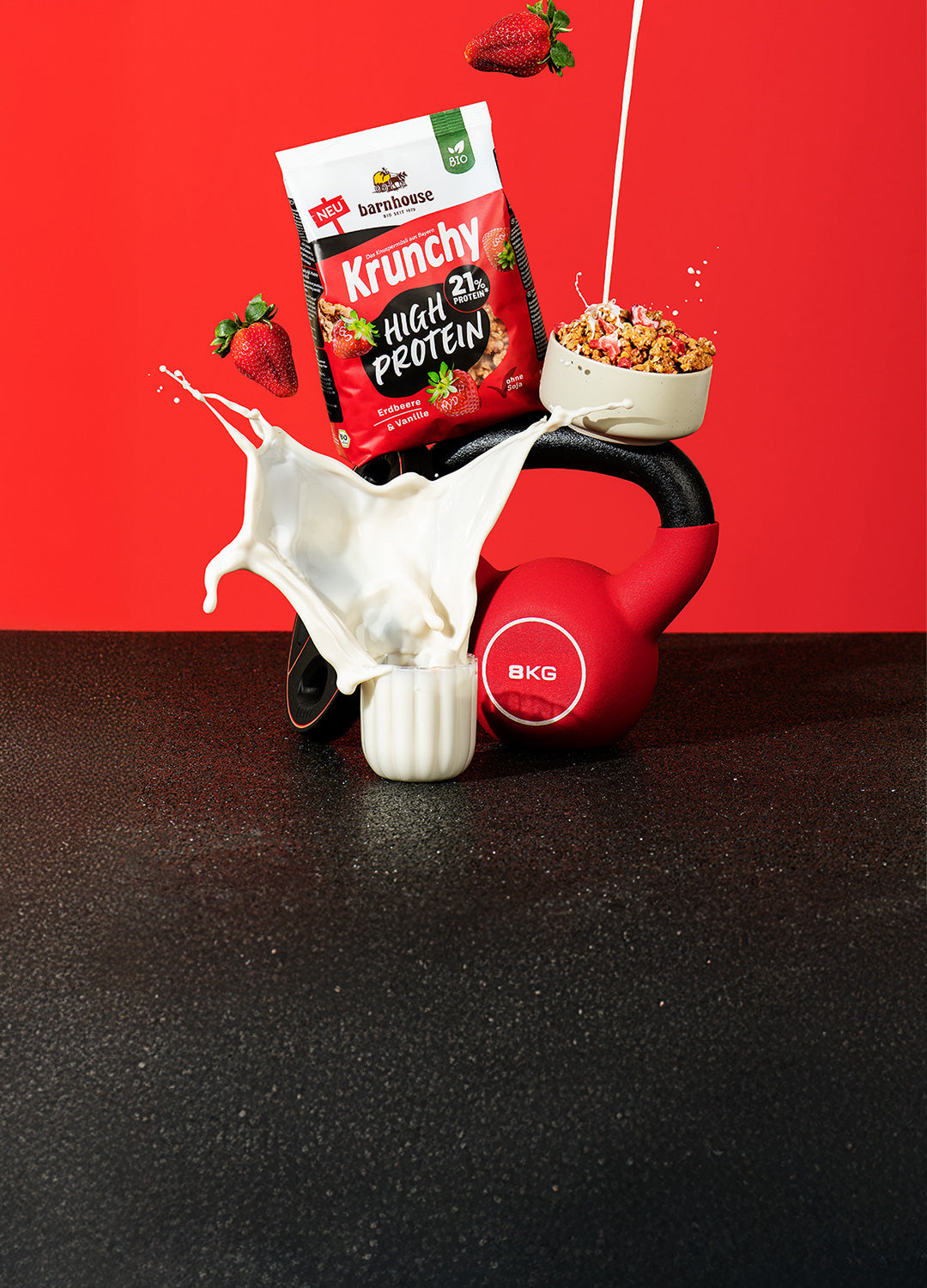 Blick von vorne auf einen Beutel Krunchy High Protein von Bio-Knuspermüsli-Hersteller Barnhouse, der auf einem Hantelgewicht und einer Kugelhantel steht. Davor steht ein Glas, aus dem Milch herausspritzt. Außerdem befindet sich noch eine Schüssel mit Krunchy High Protein auf der Kugelhantel, in die von oben ein Strahl Milch fließt. Zudem fliegen noch 2 Erdbeeren durch das Bild.
