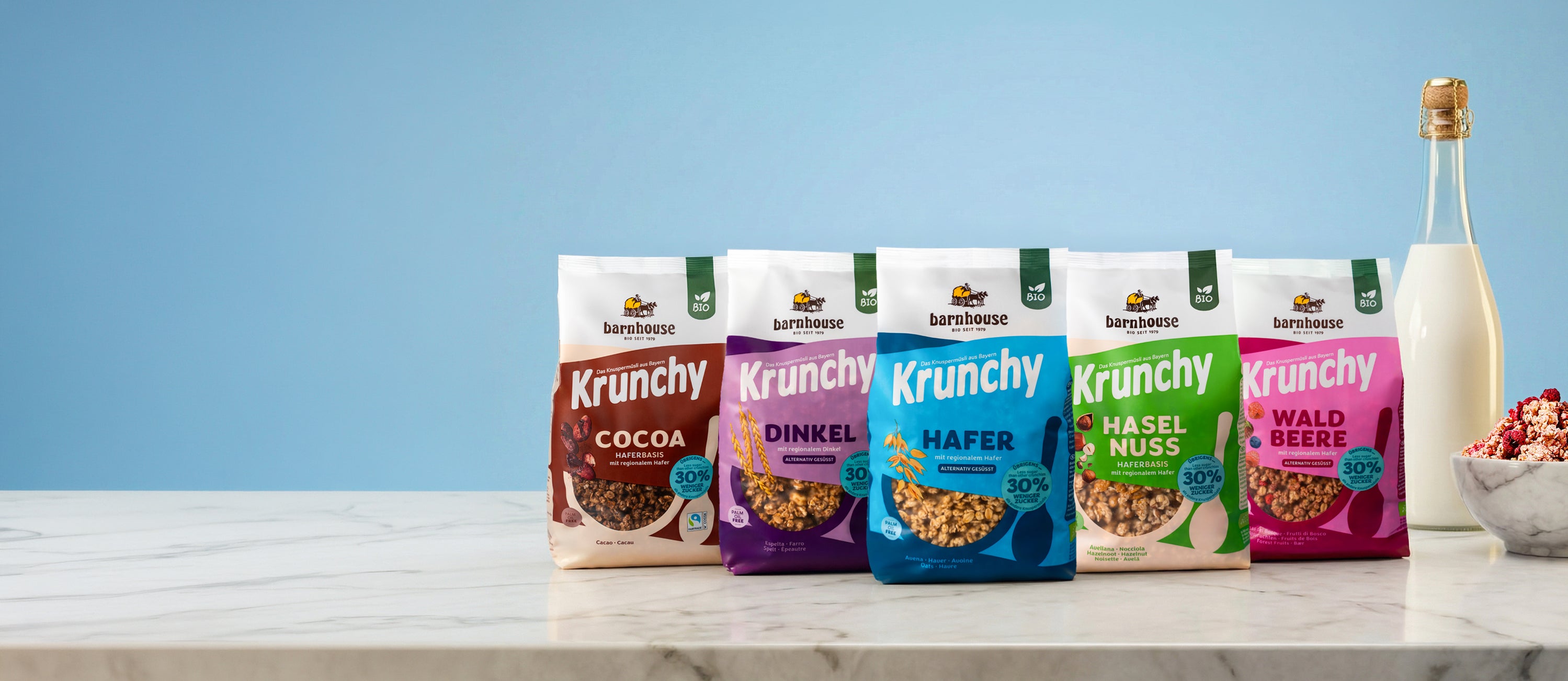 Blick von vorne auf eine Marmorplatte, auf der die fünf neuen Sorten mit 30% weniger Zucker stehen: Cocoa, Dinkel, Hafer, Haselnuss und Waldbeere. Rechts daneben eine Glasflasche mit Milch und eine Marmorschale, die mit Krunchy Waldbeere 30% weniger Zucker gefüllt ist.
