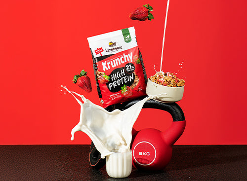 Blick von vorne auf einen Beutel Krunchy High Protein von Bio-Knuspermüsli-Hersteller Barnhouse, der auf einem Hantelgewicht und einer Kugelhantel steht. Davor steht ein Glas, aus dem Milch herausspritzt. Außerdem befindet sich noch eine Schüssel mit Krunchy High Protein auf der Kugelhantel, in die von oben ein Strahl Milch fließt. Zudem fliegen noch 2 Erdbeeren durch das Bild.