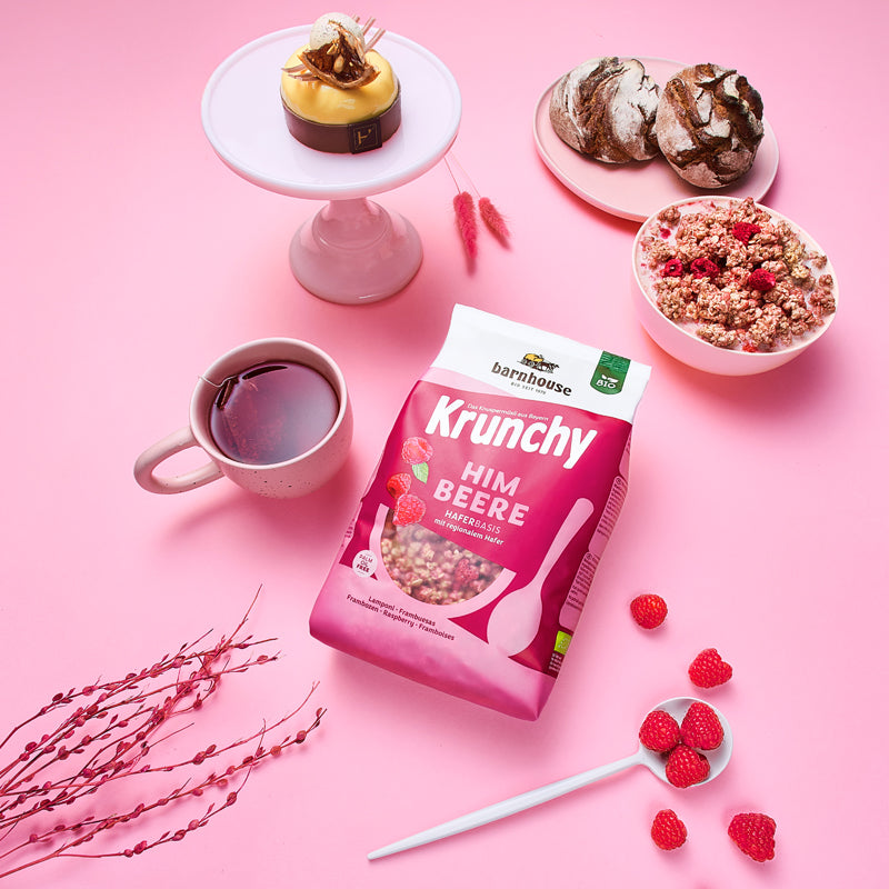 Blick von oben auf den beerenfarbigen, rosa-weißer Beutel Krunchy Himbeere von Bio-Knuspermüsli-Hersteller Barnhouse, der auf einem rosafarbenen Tisch liegt. Außenrum stehen eine Schale mit diesem Müsli und Milch, eine Tasse mit Früchtetee, ein Teller mit 2 Leib Brot, sowie ein Eierlikör-Dessert auf einem weißen Teller und ein weißer Löffel mit Himbeeren.