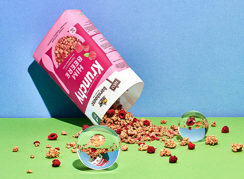 Blick von vorne auf den beerenfarbigen, rosa-weißen Beutel Krunchy Himbeere von Bio-Knuspermüsli-Hersteller Barnhouse. Der Beutel ist auf den Kopf gedreht und geöffnet, wodurch jede Menge Krunchy-Cluster und Himbeeren herausrieseln und auf den grünen Untergrund fallen. Zwischendrin liegen 2 Glaskugeln, die Elemente des Bildausschnittes spiegeln. Die Wand im Hintergrund ist blau.