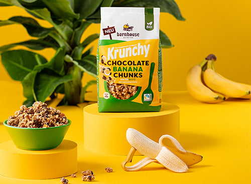 Gelber Hintergrund mit einer grünen Pflanze. Rechts daneben liegen ein paar Bananen und links ein weißer Löffel. In der Bildmitte stehen ein Beutel Krunchy Chocolate Banana Chunks von Bio-Knuspermüsli-Hersteller Barnhouse und eine grüne Schüssel mit diesem Müsli auf zwei verschiedenen gelben Sockeln. Vorne liegen noch ein paar Cluster sowie eine "Chiller"-Banane, die zur Hälfte geschält ist und einen Teil ihrer Schale als Arm nutzt, auf dem sich die restliche Banane lässig abstützt.