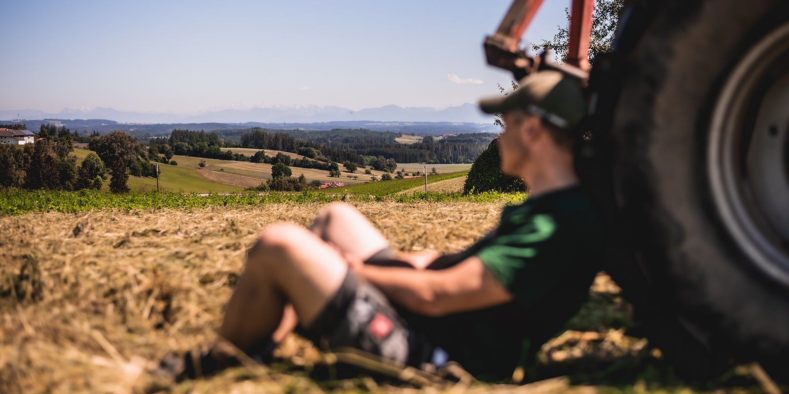 Barnhouse Bauer Sepp Heindl, der nach getaner Abend in seiner frisch gemähten Bio-Haferfeld liegt und sich am Reifen seines Traktors anlehnt. Im Hintergrund hat man eine wunderbare Aussicht auf die Landschaft in der Kirchdorfer Umgebung. Der Blick reicht sogar bis in die Berge.
