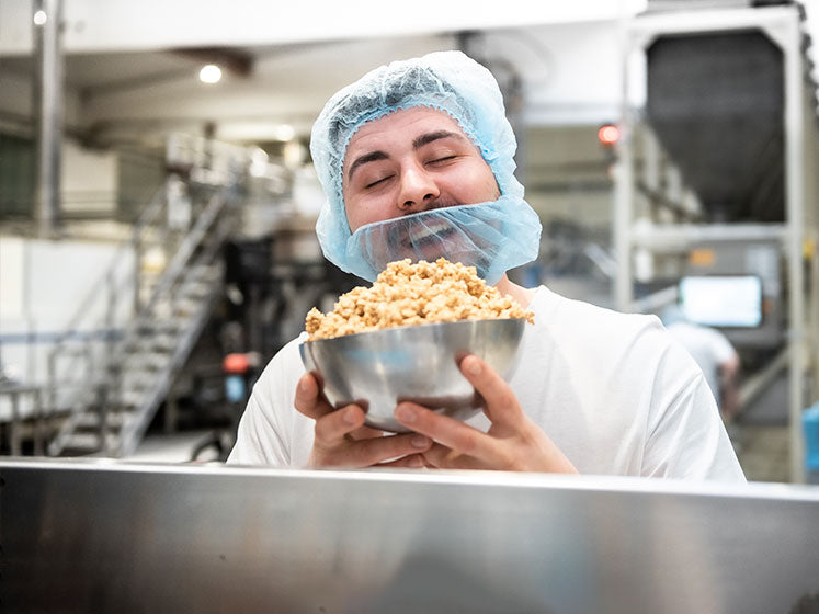Ausbilder Adrian steht mit Haube und Bartschutz in der Produktion und hält mit einem glücklichen Gesichtsausdruck und geschlossenen Augen eine Schale mit Krunchy in beiden Händen.
