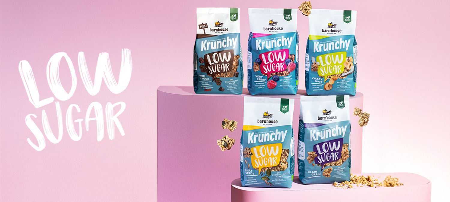 Blick von vorne auf die fünf Low Sugar Krunchys Plain Grain, Crazy Nuts, Very Berry, (No) Choco und Silly Seeds von Bio-Knuspermüsli-Hersteller Barnhouse, die auf zwei verschiedenen rosa Sockeln stehen. Außenrum fliegen Cluster. Links steht der Schriftzug "Low Sugar" vor rosa Hntergrund.