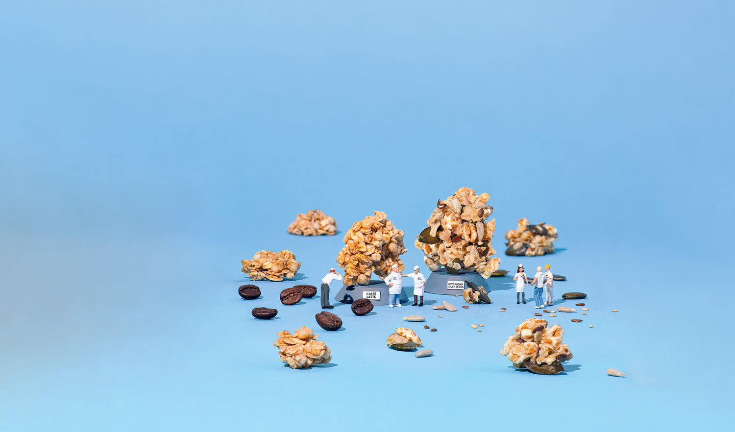 Kaffeebohnen, Kürbiskerne, Sonnenblumenkerne, Chiasamen und Leinsamen liegen gemeinsam mit Knuspermüsli-Cluster auf blauem Untergrund. In der Mitte stehen zwei kleine Podeste mit der Aufschrift "CAFFÈ LATTE" bzw. "LOS SUGAR SILLY SEEDS", auf denen der jeweilige Cluster steht. Außerdem sind sechs Männchen zu sehen, die wie Barnhouse Bäcker gekleidet sind.