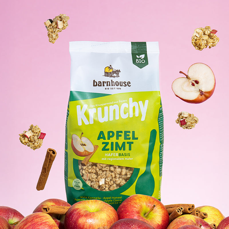 Beutel Krunchy Apfel-Zimt von Bio-Knuspermüsli-Hersteller Barnhouse, der auf einem Berg aus Äpfeln und Zimtstangen steht. Von oben fallen Knuspermüsli-Cluster mit eingebackenen Apfel-Stückchen sowie Zimtstangen und einer Apfelhälfte nach unten. Der Hintergrund ist eine Mischung aus Rosa und Weiß.
