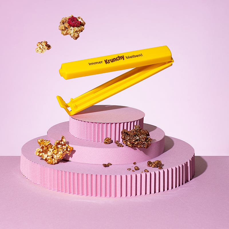 Gelber Beutelclip des Bio-Knuspermüsli-Herstellers Barnhouse mit der braunen Aufschrift "Immer Krunchy bleiben", der aufgeklappt auf einem pinken, 3-stöckigen Podest "tanzt". Außemrum schweben verschiedene Cluster.