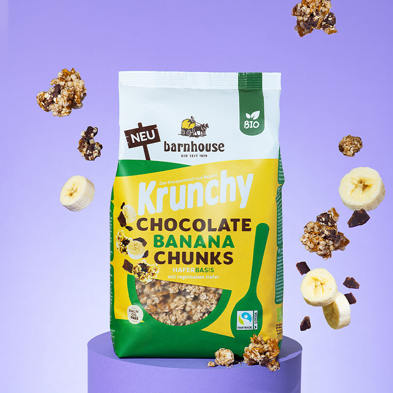 Beutel Krunchy Chocolate Banana Chunks von Bio-Knuspermüsli-Hersteller Barnhouse, der auf einem violetten, runden Socken steht. Von oben fallen Knuspermüsli-Cluster, Bananen- und Schoko-Stücke vor dem violetten Hintergrund nach unten.