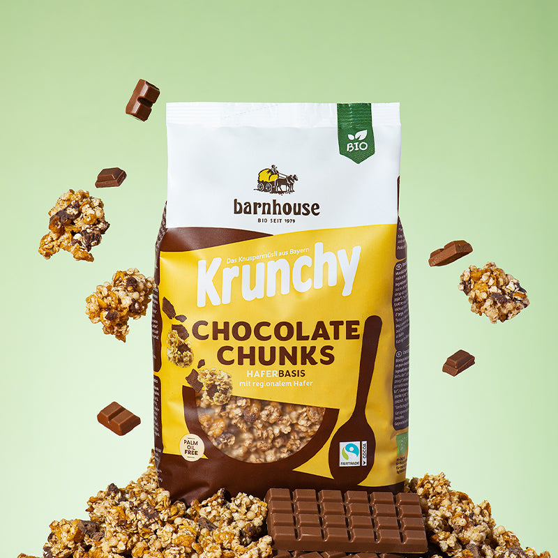 Beutel Krunchy Chocolate Chunks von Bio-Knuspermüsli-Hersteller Barnhouse, der auf einem Berg seiner Cluster sowie Schokoladebtafeln steht. Von oben fallen Knuspermüsli-Cluster  und Schoko-Stücke vor dem grün-weißen Hintergrund nach unten.