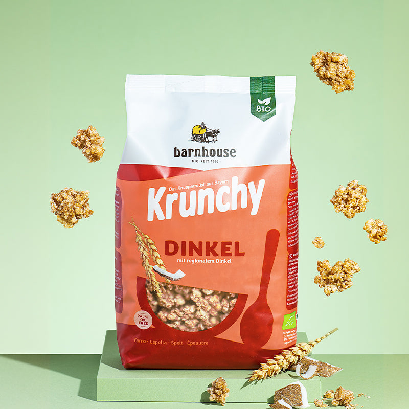 Beutel Krunchy Dinkel von Bio-Knuspermüsli-Hersteller Barnhouse, der auf einer grünen Platte steht. Von oben fallen Knuspermüsli-Cluster vor grünem Hintergrund nach unten. Um den Beutel herum liegen noch weitere Cluster, Stücke einer Kokosnuss sowie ein Dinkelhalm.