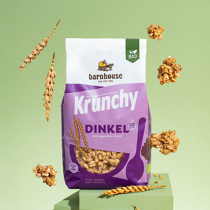Beutel Krunchy Dinkel alternativ gesüßt von Bio-Knuspermüsli-Hersteller Barnhouse, der auf 2 grünen Treppenstufen steht. Von oben fallen Knuspermüsli-Cluster sowie Dinkelhalme vor einem grünen Hintergrund nach unten.