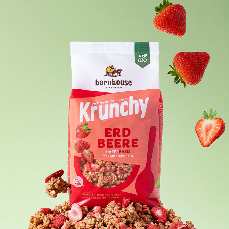 Beutel Krunchy Erdbeere von Bio-Knuspermüsli-Hersteller Barnhouse, der auf einem Berg aus Müsli-Clustern und Erdbeerscheiben steht. Von oben fallen rote, saftige Erdbeeren nach unten.