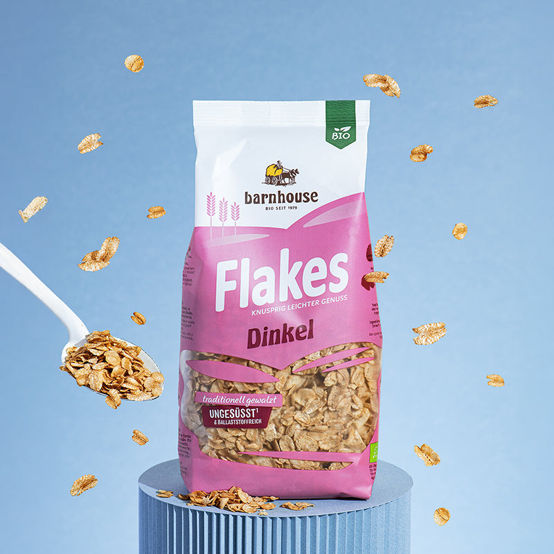 Beutel Flakes Dinkel vom Bio-Knuspermüsli-Hersteller Barnhouse, der vor blauem Hintergrund auf einem blauen Sockel steht. Im ganzen Bild schweben Dinkelflakes rum. Links ist ein weißer Löffel mit weiteren Dinkelflakes zu sehen.