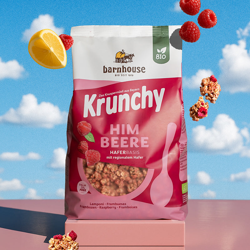 Beerenfarbiger, rosa-weißer Beutel Krunchy Himbeere von Bio-Knuspermüsli-Hersteller Barnhouse auf einem eckigen Sockel. Von oben fallen Knuspermüsli-Cluster, Himbeeren & 1 Zitronen-Viertel vor einem Hintergrund mit blauem Himmel und Wolken nach unten.