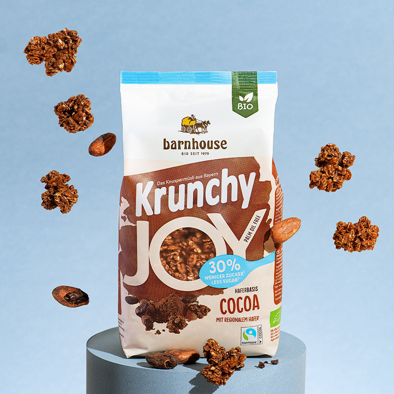 Beutel Krunchy Joy Cocoa von Bio-Knuspermüsli-Hersteller Barnhouse, der auf einem runden, blauen Sockel steht. Von oben fallen Knuspermüsli-Cluster sowie Kakaobohnen vor einem blauen Hintergrund nach unten.
