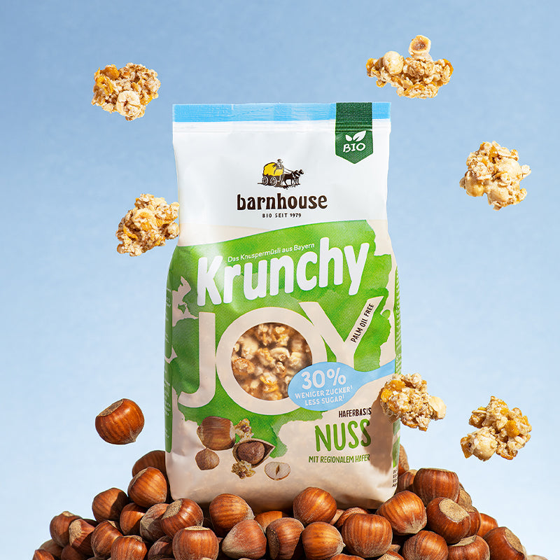 Grün-weißer Beutel Krunchy Joy Nuss von Bio-Knuspermüsli-Hersteller Barnhouse, der auf einem Berg aus Haselnüssen steht. Von oben fallen Knuspermüsli-Cluster vor einem blauen Hintergrund nach unten.