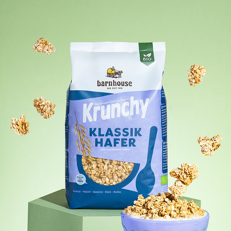 Blau-weißer Beutel Krunchy Klassiker Hafer von Bio-Knuspermüsli-Hersteller Barnhouse, der auf einem eckigen, grünen Sockel steht. Von oben fallen Knuspermüsli-Cluster vor einem grünen Hintergrund nach unten. Vorne rechts steht eine Müslischale im Blau des Beutels, in der etwas Milch und das Hafer-Müsli eingefüllt sind. 2 große Cluster fliegen gerade von oben in die Schale.