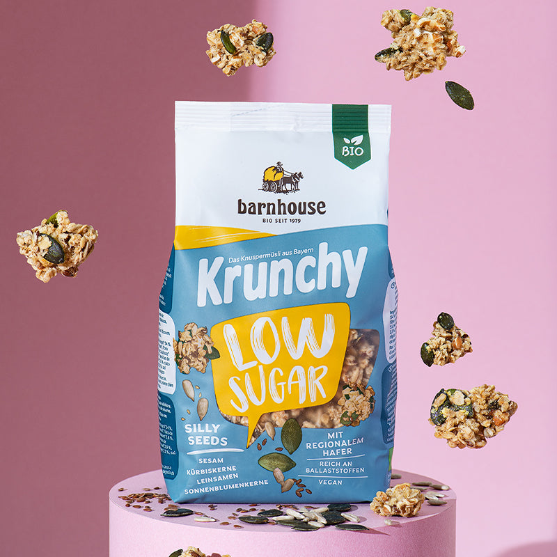 Gelb-weiß-blauer Beutel Krunchy Low Sugar Silly Seeds von Bio-Knuspermüsli-Hersteller Barnhouse, der auf einem runden, rosa Sockel steht. Von oben fallen Knuspermüsli-Cluster und ein Kürbiskern vor einem rosa Hintergrund nach unten. Auf dem Sockel liegen neben einem Cluster auch Kürbiskerne, Leinsamen und Sonnenblumenkerne - die Zutaten des zuckerarmen Müslis.