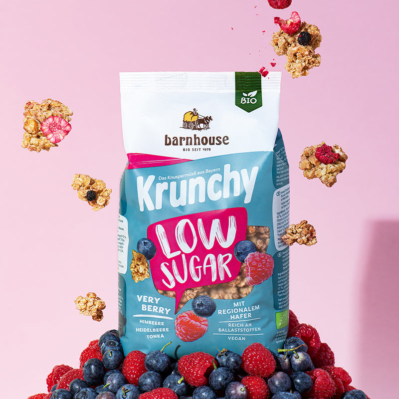 Pink-weiß-blauer Beutel Krunchy Low Sugar Very Berry von Bio-Knuspermüsli-Hersteller Barnhouse, der auf einem Berg aus Himbeeren und Heidelbeeren steht. Von oben fallen Knuspermüsli-Cluster mit den Beeren vor einem rosa Hintergrund nach unten.