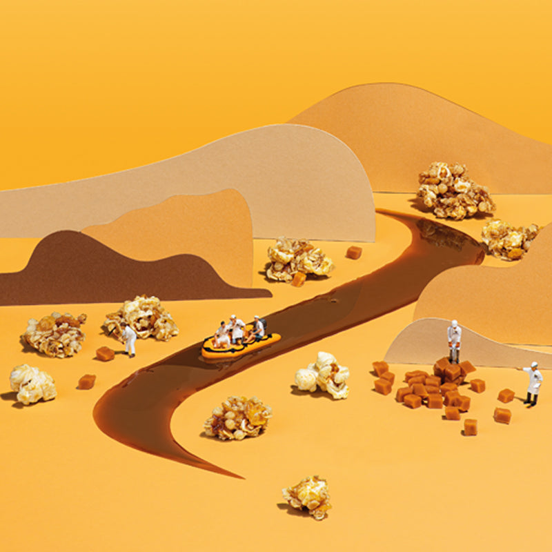 Bild-Animation, bei der in der Mitte ein Fluss aus Karamell fließt. Darauf fährt ein gelbes Boot mit 3 Barnhouse Bäckern in Männchenform. Links und rechts beschäftigen sich 3 weitere Barnhouse Bäcker mit Karamell-Stückchen. Die Kulisse zeigt außerdem noch Popcorn und Müsli-Cluster sowie wellenförmige Kartons, die Berge symbolisieren.