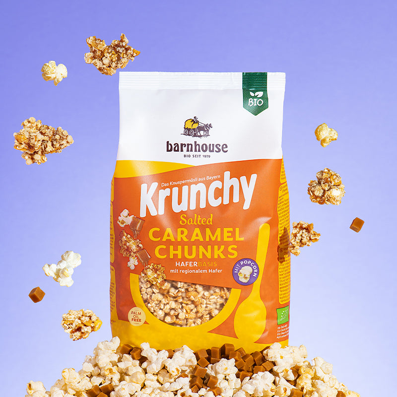 Gelb-weiß-oranger Beutel Krunchy Salted Caramel Chunks von Bio-Knuspermüsli-Hersteller Barnhouse, der auf einem Berg aus Popcorn und Karamell-Stückchen steht. Von oben fallen Knuspermüsli-Cluster, Popcorn und Karamell-Stückchen vor einem blauen Hintergrund nach unten.