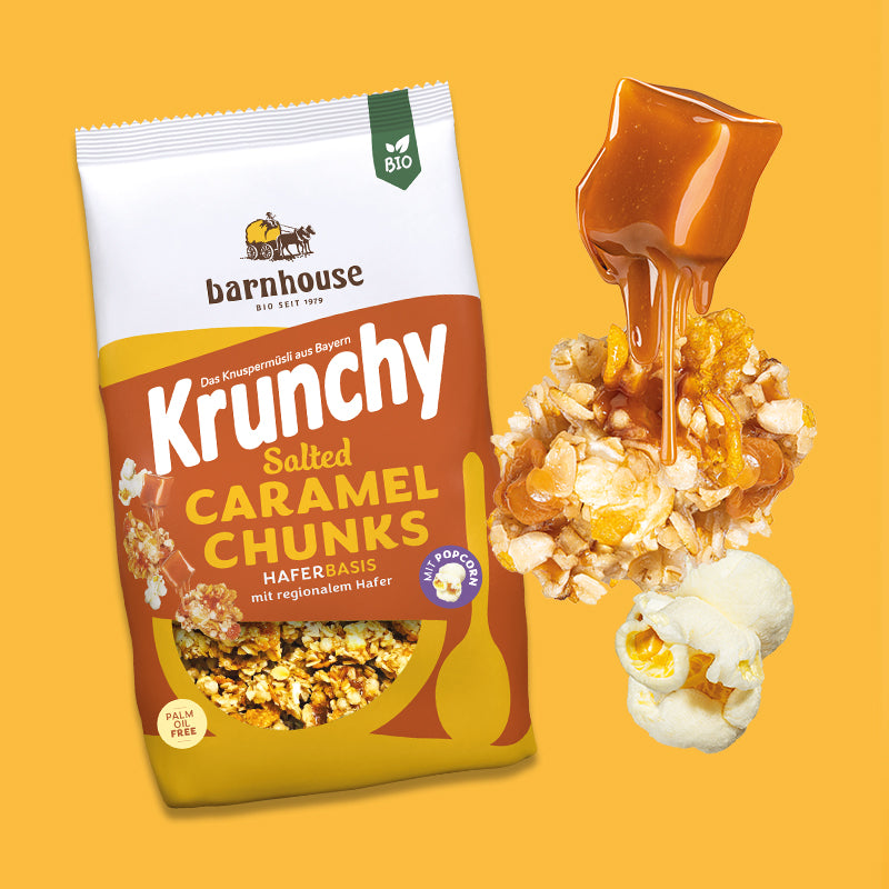Gelb-weiß-oranger Beutel Krunchy Salted Caramel Chunks von Bio-Knuspermüsli-Hersteller Barnhouse, der vor gelbem Hintergrund steht. Rechts daneben fließt ein Karamell-Stück langsam in ein Müsli-Cluster ein, während darunter ein Popcorn hängt.