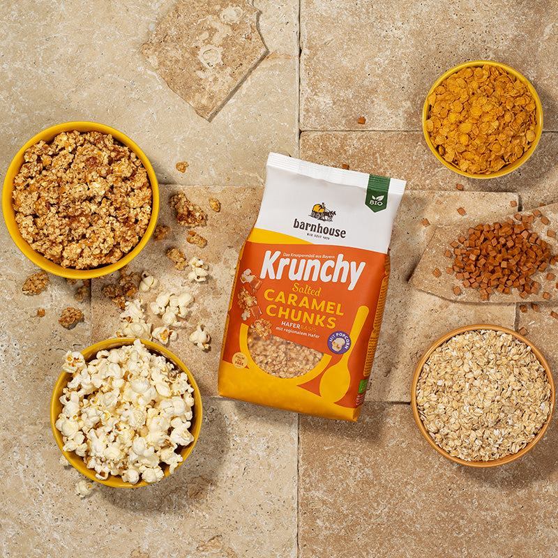 Blick von oben auf einen gelb-weiß-orangen Beutel Krunchy Salted Caramel Chunks von Bio-Knuspermüsli-Hersteller Barnhouse, der auf Fliesen liegt. Daneben liegen Karamell-Stückchen, Popcorn und Müsli-Cluster. Außerdem stehen noch 3 gelbe Schalen mit den Zutaten Popcorn, Cornflakes und Haferflocken außenrum. Die vierte gelbe Schale ist voll mit dem Müsli.