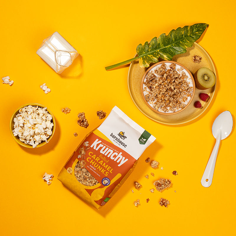 Blick von oben auf einen gelb-weiß-orangen Beutel Krunchy Salted Caramel Chunk von Bio-Knuspermüsli-Hersteller Barnhouse auf gelbem Untergrund. Außenrum sind Popcorn und Müsli-Cluster verteilt. Während links eine gelbe Schale mit Milch sowie eine Karaffe mit Milch stehen, liegt auf der rechten Seite ein weißer Löffel. Oben rechts steht eine orange Schüssel mit Milch und dem Karamell-Müsli. Darunter ein Unterteller, auf dem noch ein grünes Blatt, ein Cluster sowie zwei Himbeeren und eine halbe Kiwi liegen.