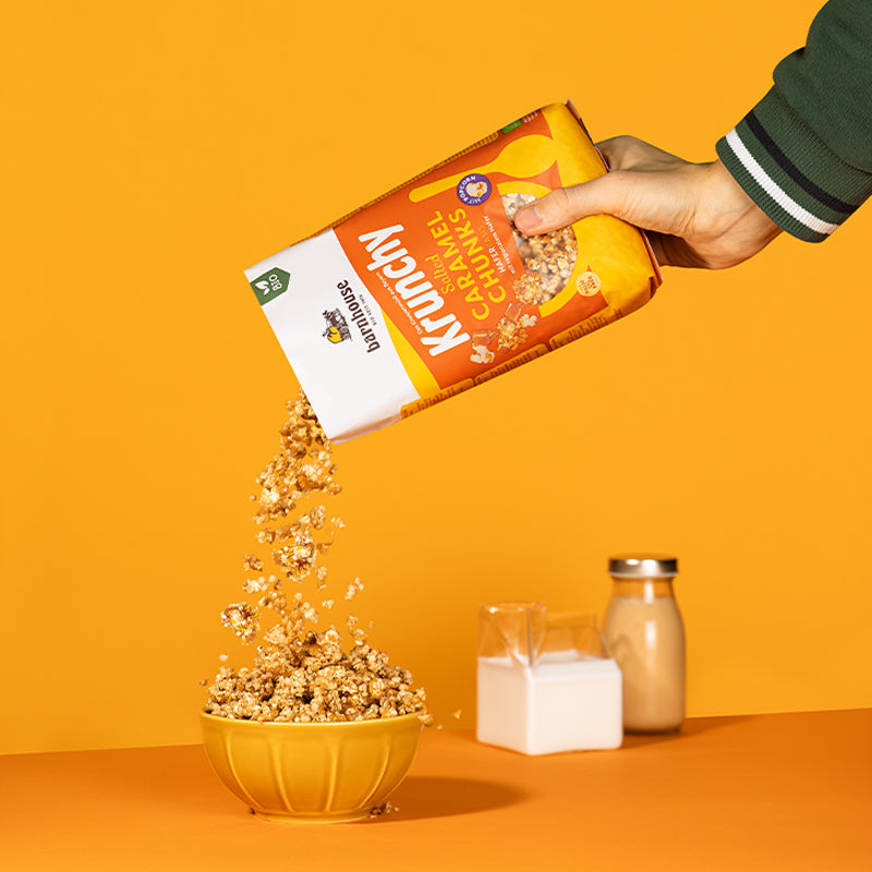 Blick von vorne, wie eine rechte Hand mit grünem Jackenärmel Krunchy Salted Caramel Chunks von Bio-Knuspermüsli-Hersteller Barnhouse aus dem gelb-weiß-orangen Beutel in eine orange Schale füllt. Während der Boden orange ist, hat die Wand eine gelbe Farbe. Im Hintergrund stehen noch ein Gefäß mit Milch und eine Flasche mit Milchalternative.