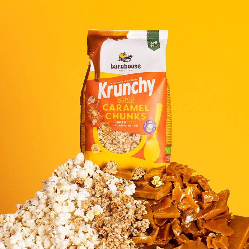 Gelb-weiß-oranger Beutel Krunchy Salted Caramel Chunks von Bio-Knuspermüsli-Hersteller Barnhouse, der auf einem Berg aus Popcorn, Müsli-Cluster und Karamell-Stückchen steht. Vom oberen, linken Eck des Beutels fließt Karamell schön nach unten. Der Hintergrund ist gelb.