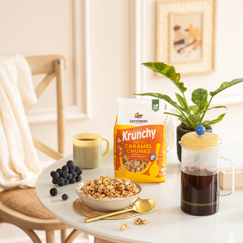 Blick von vorne auf einen hellen Frühstückstisch. Im Hintergrund ein Bild sowie ein Stuhl mit Handtuch. Auf dem Tisch dreht es sich ums Frühstück. Vor einer grünen Pflanze sorgt der gelb-weiß-orange Beutel Krunchy Salted Caramel Chunks von Bio-Knuspermüsli-Hersteller Barnhouse für einen Blickfang. Daneben stehen eine Tasse sowie eine Kanne mit Kaffee und Heidelbeeren. Vorne liegt ein geflochtenes Set mit einem goldenen Löffel und einer weißen Schale, befüllt mit Milch und dem Karamell-Müsli.