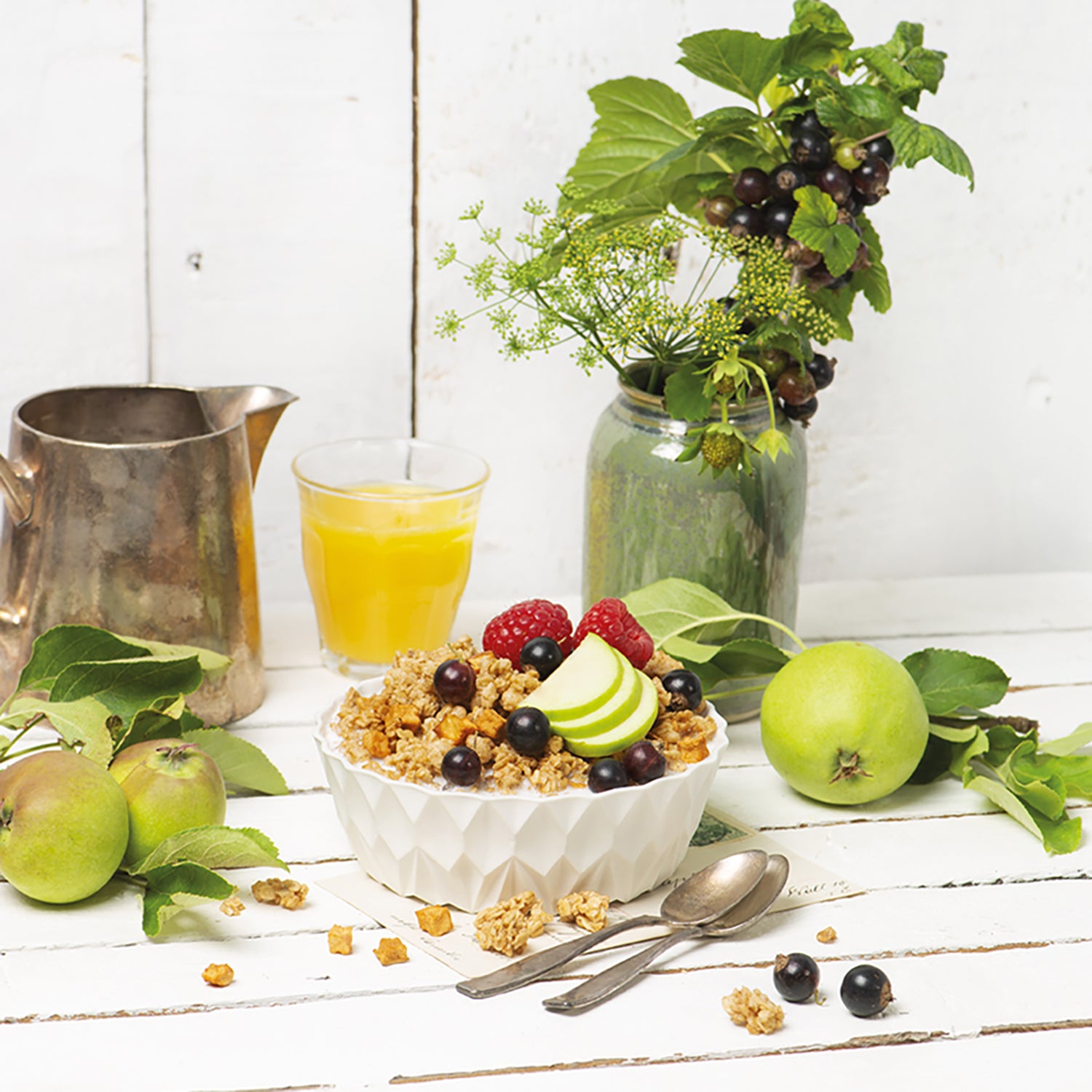 Ein sehr schön dekorierter, weißer Holztisch vor einer weißen Holzwand. Das Hauptfokus liegt auf einer edlen, weißen Schüssel, die mit Krunchy Apfel-Zimt von Bio-Knuspermüsli-Hersteller Barnhouse gefüllt ist. Als Topping liegen Himbeeren, Apfelscheiben und Johannisbeeren auf dem Müsli. Außerdem liegen noch ein paar Cluster, Johannisbeeren Apfelstückchen sowie Äpfel mit Blättern auf dem Tisch. Zudem sieht man einen Strauch aus Pflanzen und Johannisbeeren sowie ein Glas mit Orangensaft und eine Blechkanne.