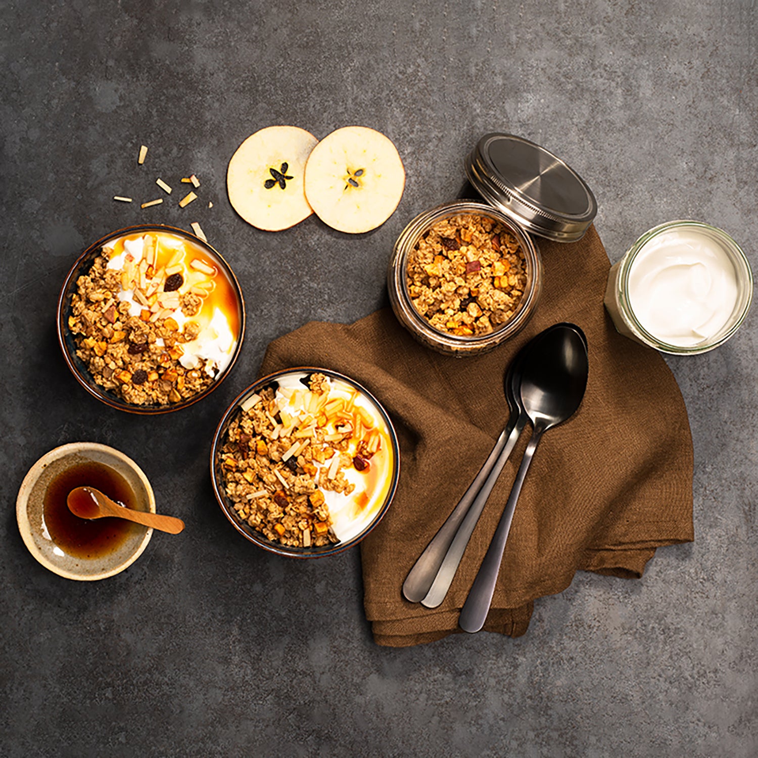 Dunkler Tisch mit einem braunen Geschirrtuch, auf dem 3 Löffel liegen. Daneben befindet sich ein durchsichtiges Gefäß mit Krunchy Apfel-Zimt von Bio-Knuspermüsli-Hersteller Barnhouse. Zudem 2 Schüsseln, die jeweils mit Krunchy Apfel-Zimt, Joghurt und Apfeldicksaft befüllt sind. Zudem stehen noch 2 Apfelscheiben, eine Schüssel mit Apfeldicksaft und Holzlöffel sowie eine Glas mit Joghurt aus dem Tisch.